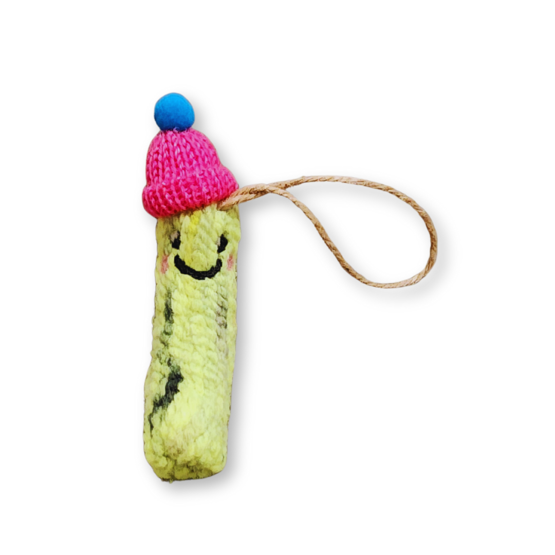 A light green felted pickle ornament with a pink knit hat topped with a blue pompom, showcasing its happy expression and a jute string hanger.