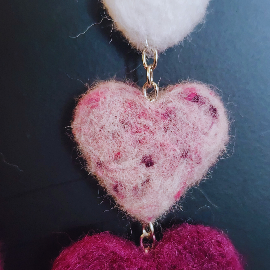 A close-up of a handmade needle-felted heart chain earring, showing a light pink felt heart with darker pink flecks, connected by a silver-toned chain to a white felt heart above it.