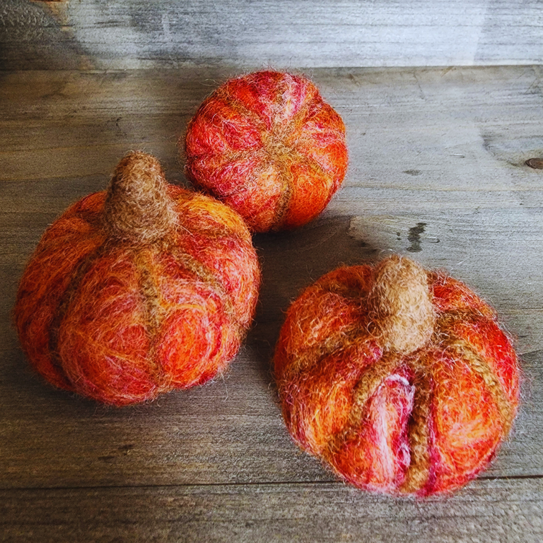 Three handmade felted pumpkins in shades of orange and red with brown felted stems, arranged in a triangular pattern on a wooden surface.
