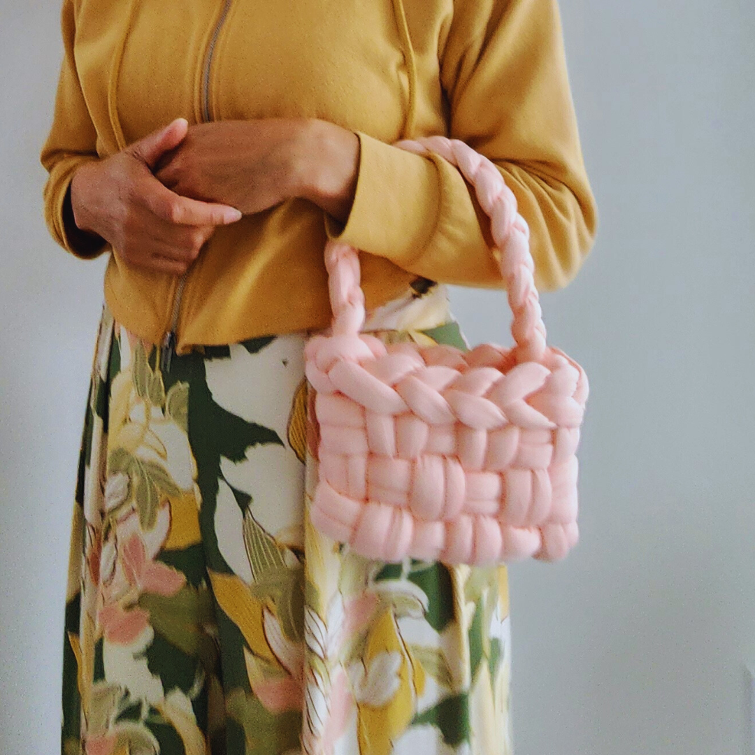 A person wearing a yellow top and a floral skirt holds a small handmade basket weave handbag in light coral pink with both hands, positioned in front of their torso.