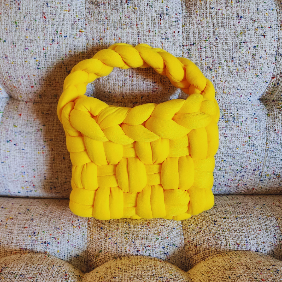 A small handmade basket weave handbag in vibrant yellow, with a thick braided handle, sitting upright on a multicolored speckled fabric surface.