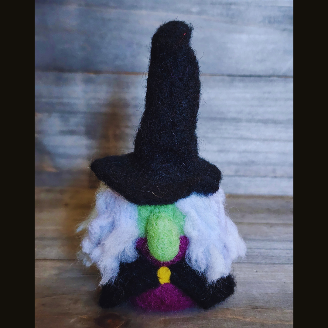 A handmade needle-felted witch figurine with a black pointy hat, a bright green face, a purple body, a yellow buckle, and fluffy white hair, standing centered against a wooden background.