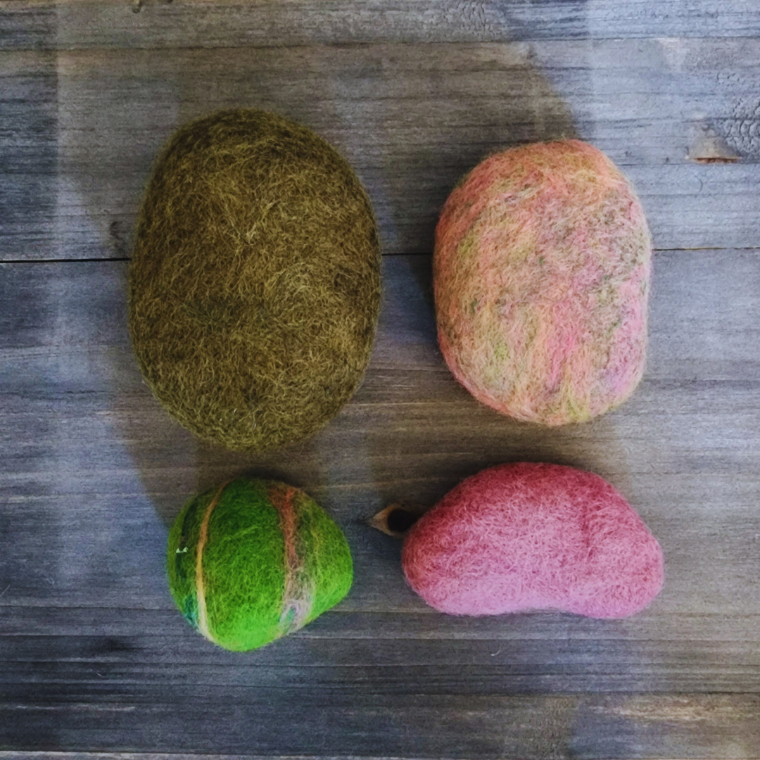 Four handmade wet-felted Zen Stones in earthy and pastel tones arranged unstacked on a wooden surface: a large olive green, a mottled pink and light green, a vibrant lime green with stripes, and a solid light pink.