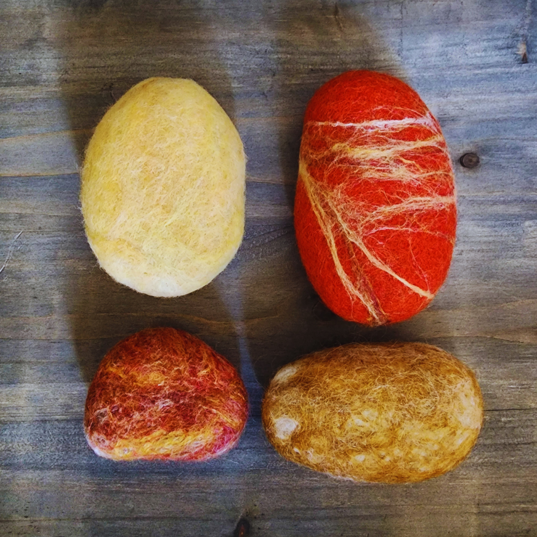 Four handmade wet-felted Zen Stones in shades of yellow, orange, and red, arranged unstacked on a wooden surface. This includes a solid light yellow, a vibrant red-orange with yellow and white streaks, a small mottled orange/red, and a golden brown.