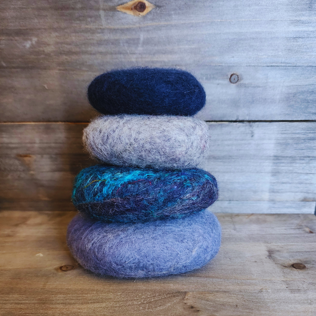 Four handmade wet-felted Zen Stones stacked vertically, featuring shades of light blue/lavender, dark blue with turquoise accents, light gray, and deep navy blue, on a rustic wooden background.