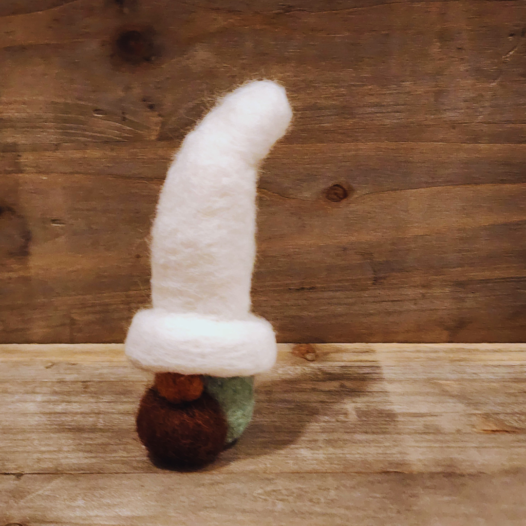 A handmade needle-felted gnome with an aqua body, a tall white hat, a brown mustache, and a light-colored nose, viewed from a side angle against a rustic wooden background.