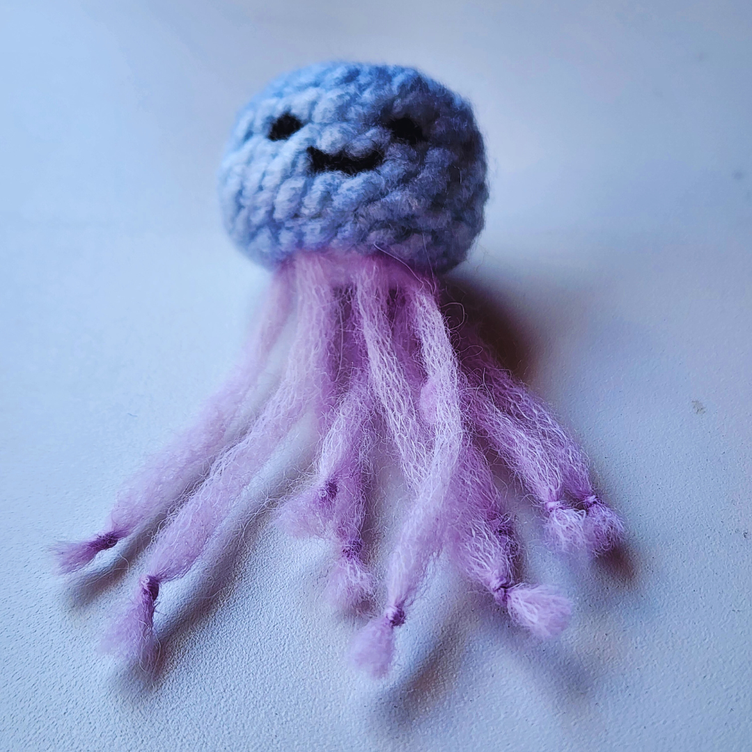 A handmade jellyfish bag charm with a light purple head and a stitched black smiley face. It has fluffy, light purple tentacles and is shown on a white background.