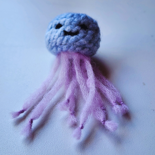 A handmade jellyfish bag charm with a light purple head and a stitched black smiley face. It has fluffy, light purple tentacles and is shown on a white background.