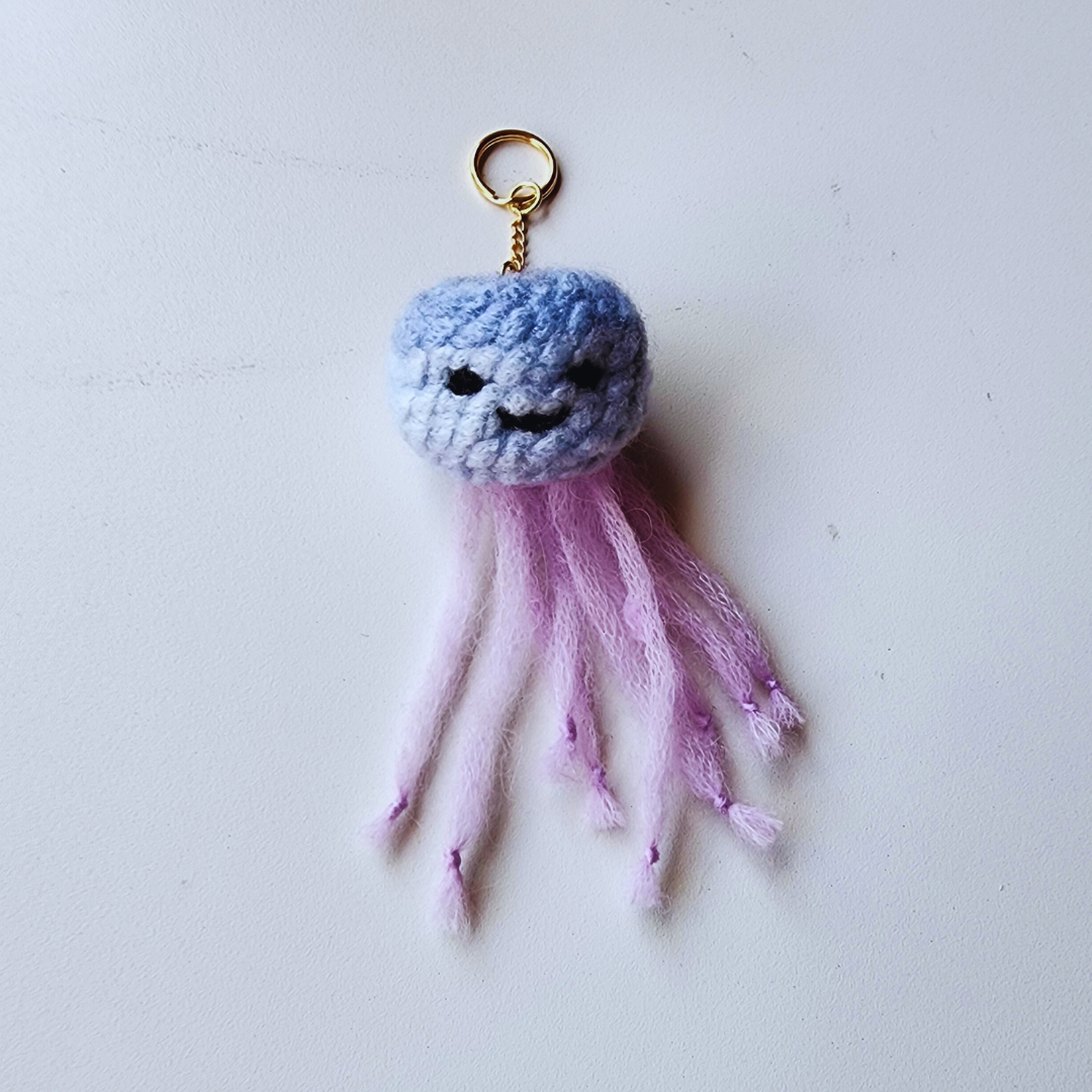 A handmade jellyfish charm is shown from the underside, revealing a round, pink and gray body with bright green rope-like tentacles and a silver clip-on keychain.