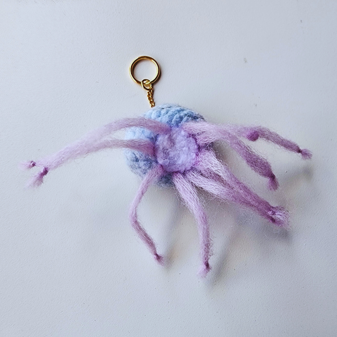 A handmade jellyfish bag charm is shown from the back, with its light purple tentacles spread out and a gold keychain ring attached.