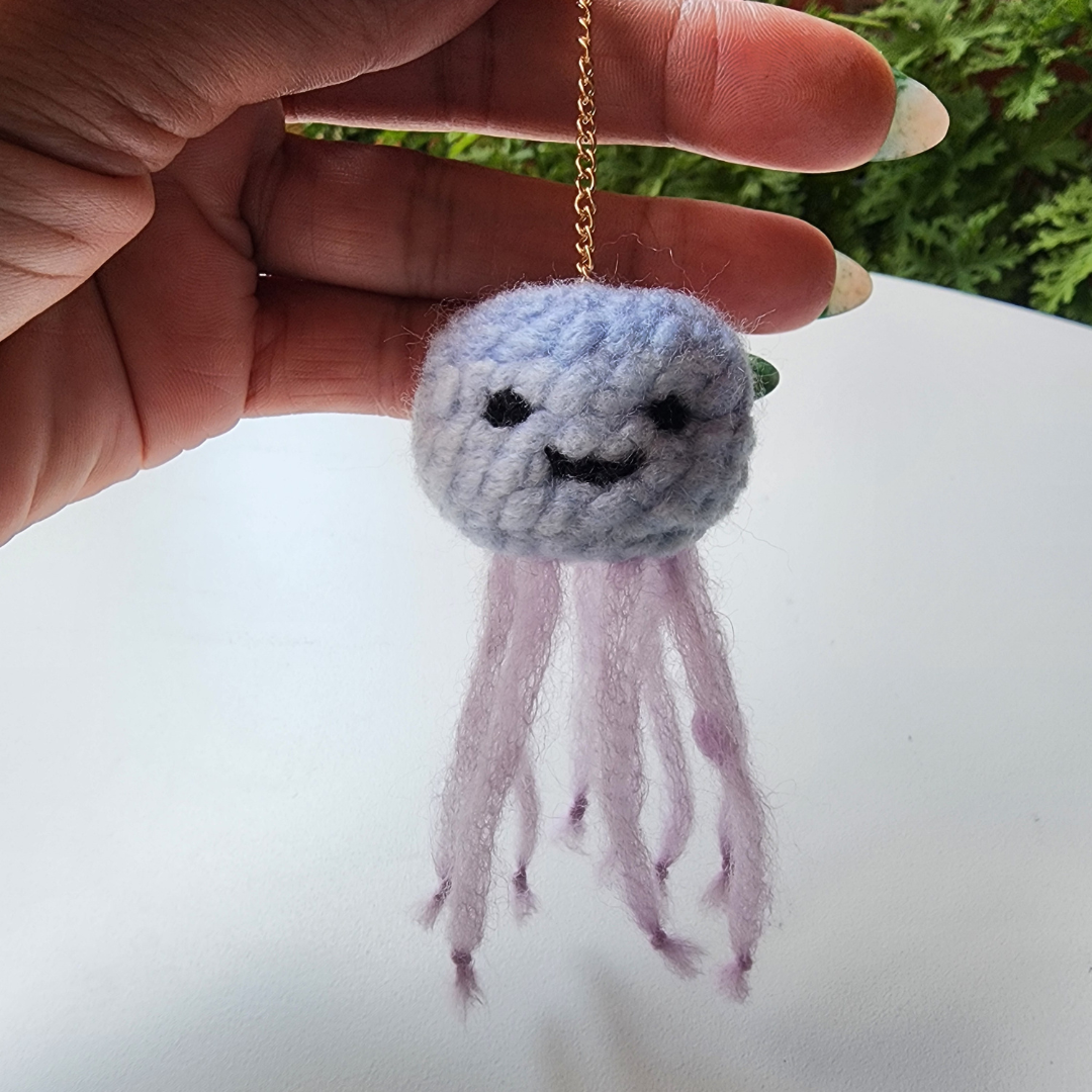 A hand holds a handmade jellyfish bag charm, showing its light purple head with an embroidered smile and matching tentacles. A gold keychain ring is visible at the top.