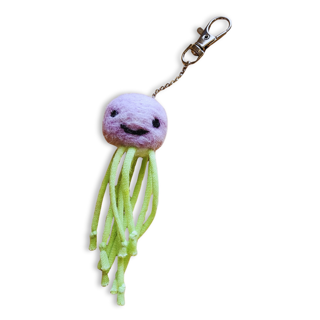 A handmade jellyfish bag charm with a pink head and a stitched black smiley face, from which hangs bright green rope-like tentacles and a silver clip-on keychain.