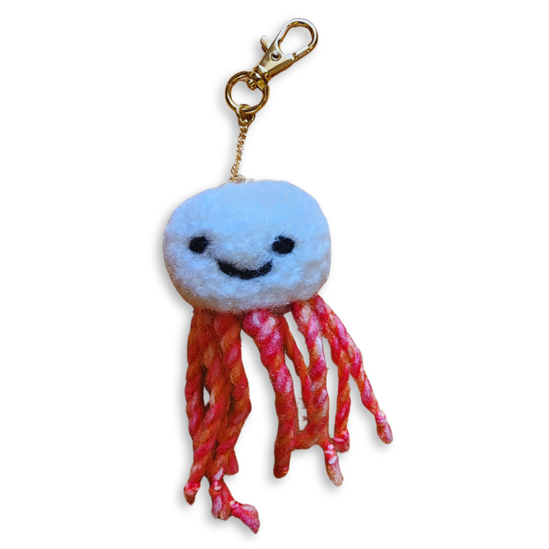 A handmade jellyfish bag charm with a white head and a stitched black smiley face. From its body hang orange and red mixed rope-like tentacles with a gold clip-on keychain.