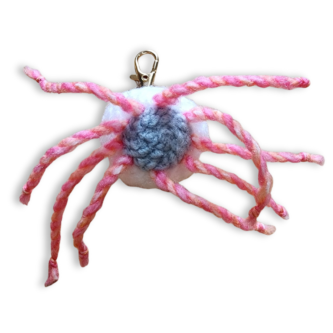 A handmade jellyfish bag charm is shown from the underside, revealing a round body with pink and gray colors, from which hang thick, bright pink rope-like tentacles. It has a gold clip-on keychain.