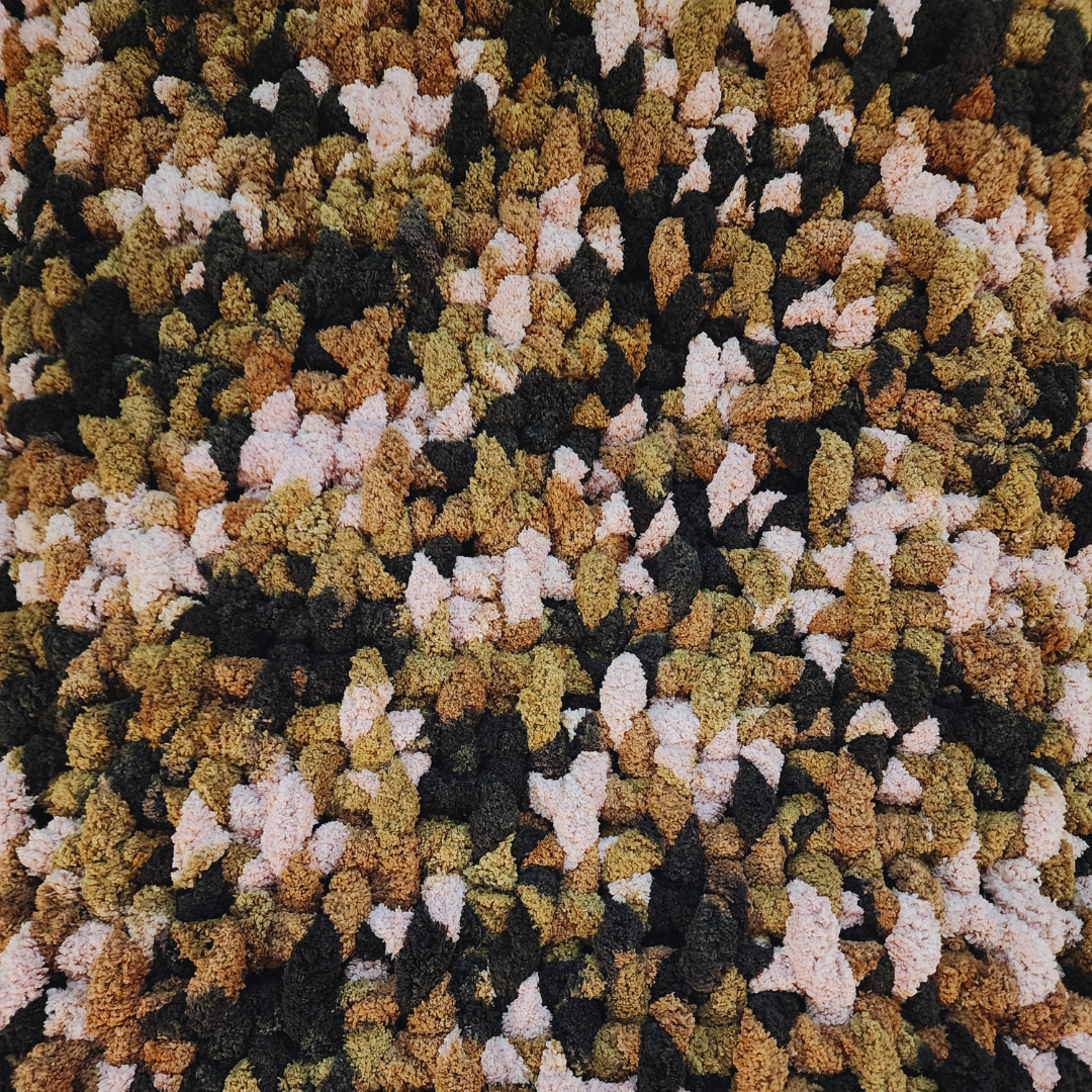 A close-up overhead view of a handmade chunky knit blanket with a basket weave stitch, showing its textured surface and multicolor pattern in shades of brown, pink, and black.