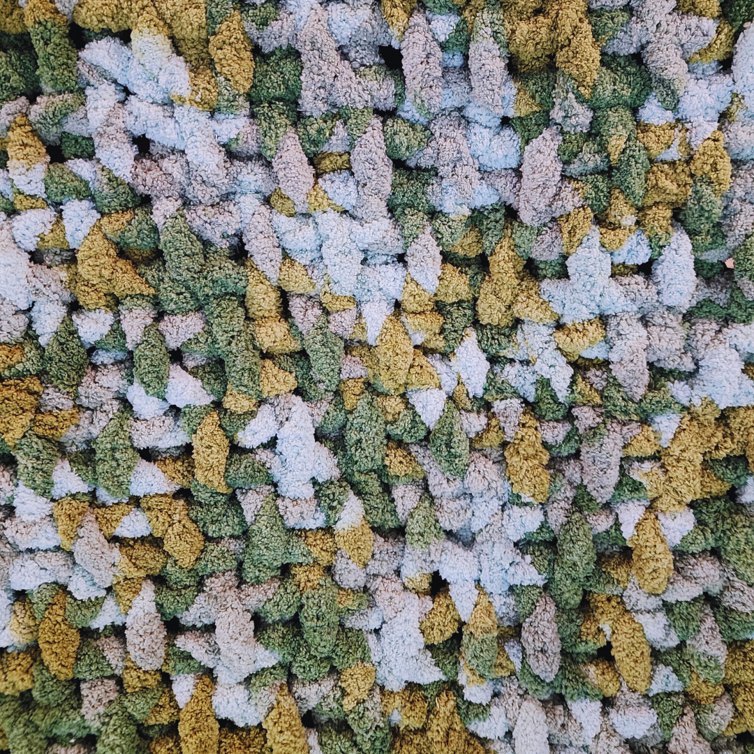 A close-up overhead view of a handmade chunky knit blanket with a basket weave stitch, showing its textured surface and multicolor pattern in shades of green, yellow, and light gray.
