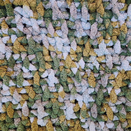 A close-up overhead view of a handmade chunky knit blanket with a basket weave stitch, showing its textured surface and multicolor pattern in shades of green, yellow, and light gray.