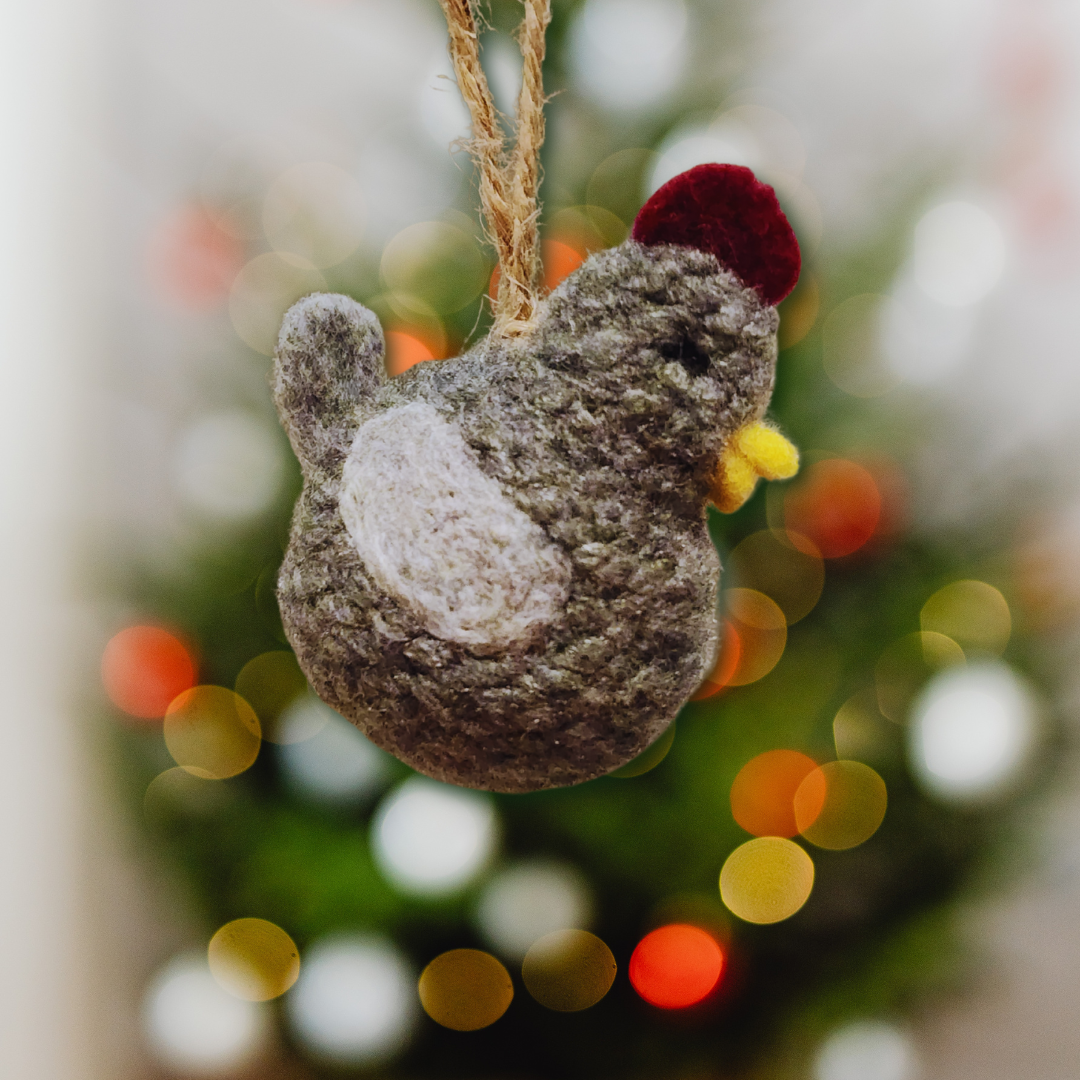 A handmade Grey chicken bobble ornament, featuring a white wing patch and red comb, hanging against a blurred Christmas tree background.
