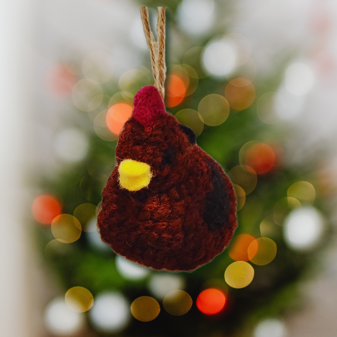 A plump, handmade Rhode Island Red chicken bobble ornament, featuring a red comb and yellow beak, hanging in front of a blurred Christmas tree background with warm lights.
