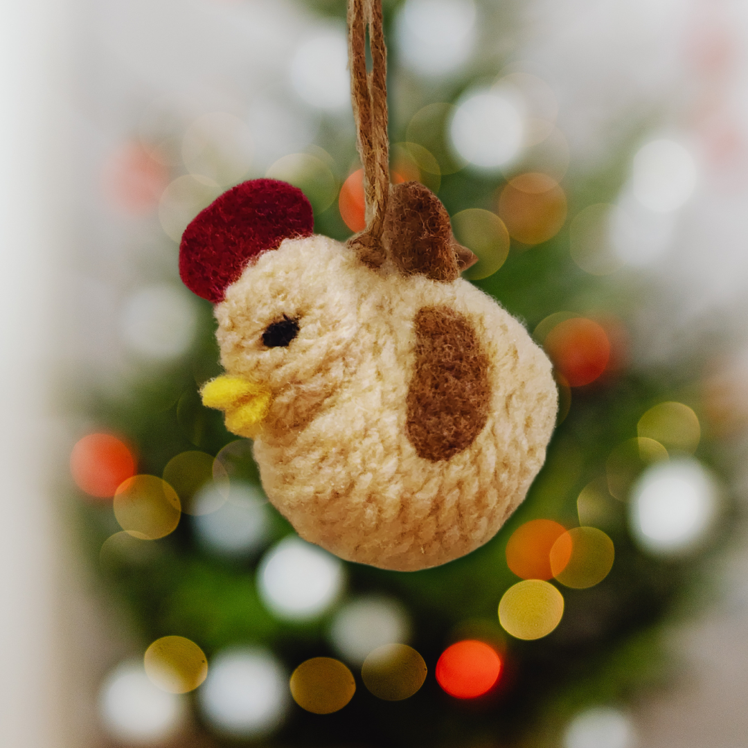 A cheerful, handmade Tan chicken bobble ornament, featuring a brown speckled patch and a yellow beak, hanging against a blurred Christmas tree background.