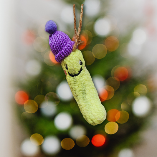 A bright green, needle-felted Christmas Pickle ornament with a smiling face and rosy cheeks, wearing a purple knit winter hat with a pompom, hanging against a blurred Christmas tree background.