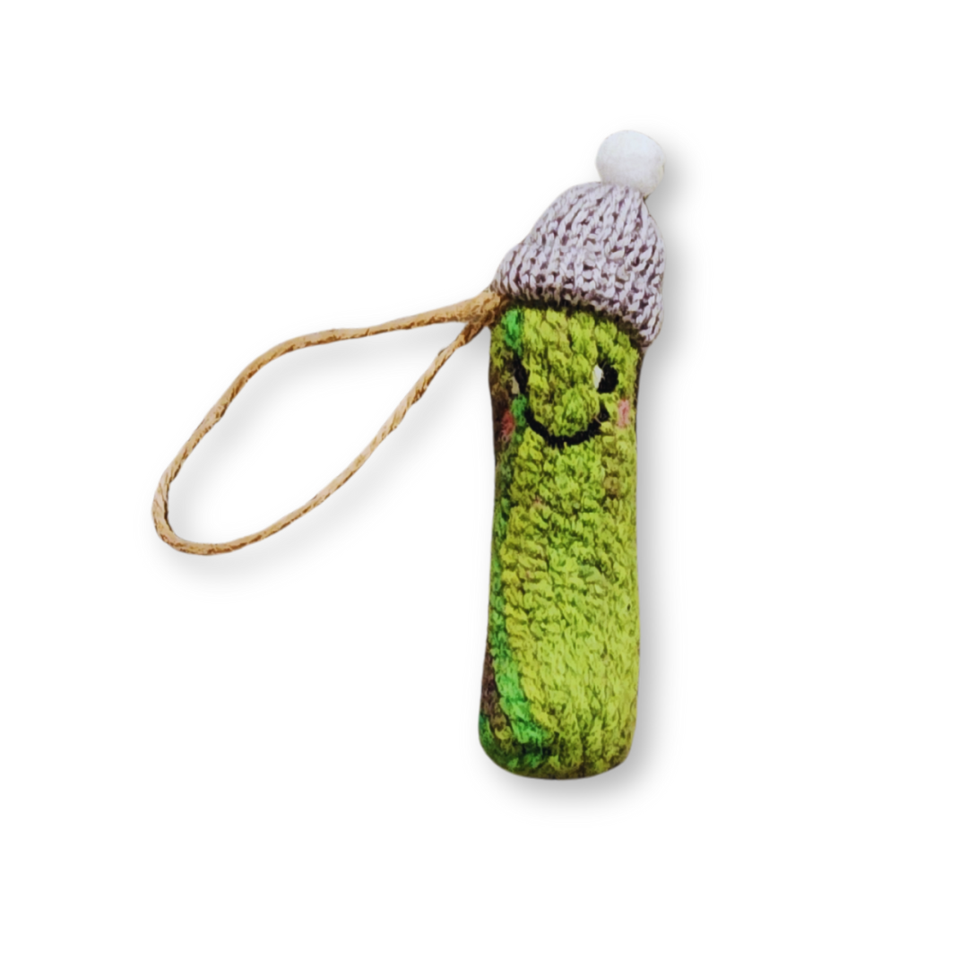 A fuzzy green felted pickle ornament wearing a light grey knit winter hat with a white pompom, hanging from a jute string.