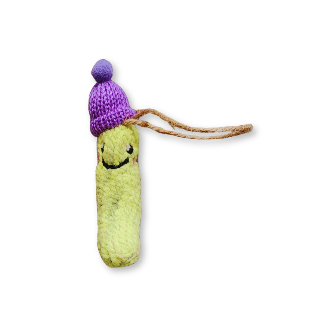 A light green felted pickle ornament with a smiling expression, wearing a bright purple knit hat with a matching pompom, and hanging from a jute string.