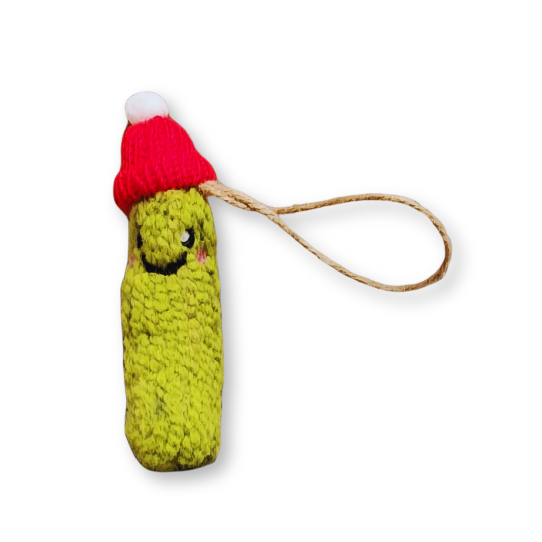 A light green felted pickle ornament with a smiling face, wearing a festive red knit Santa hat with a white pompom and hanging from a jute string.