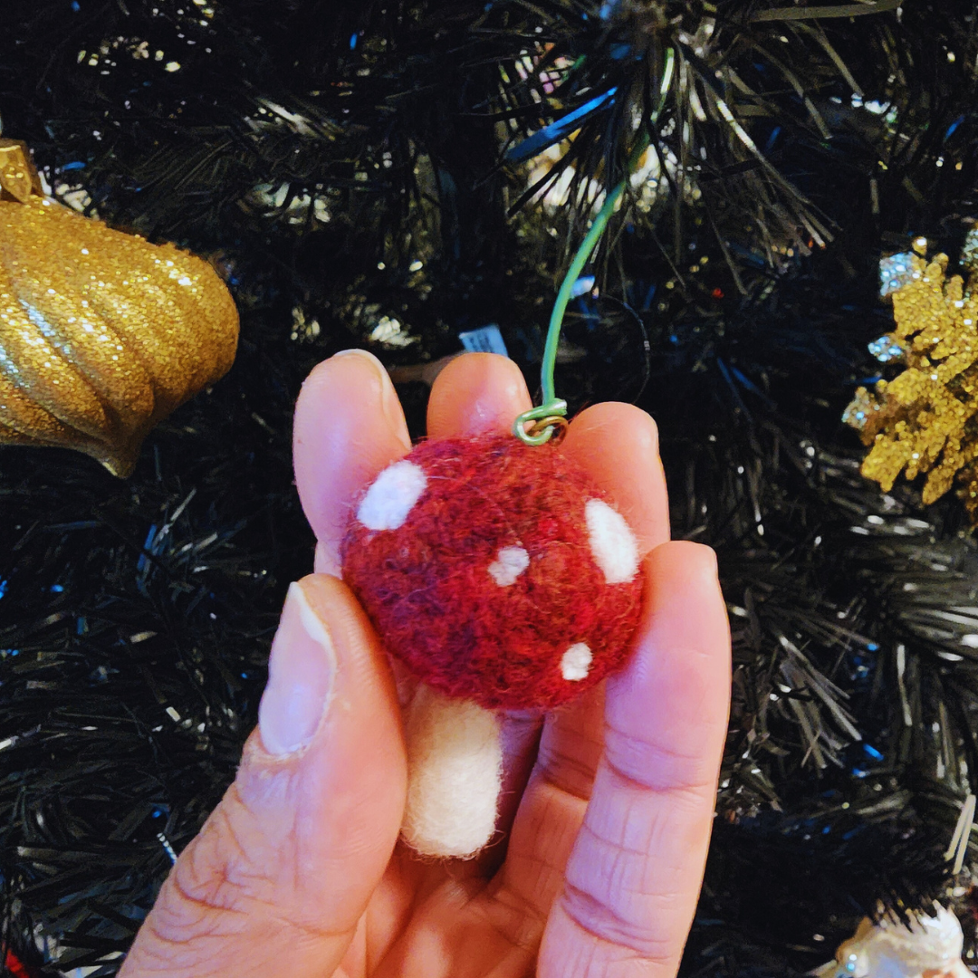 A person's hand holds a small, round needle-felted toadstool ornament (red cap with white spots, white stem) against the background of a dark Christmas tree adorned with gold ornaments. A green wire loop is attached for hanging.
