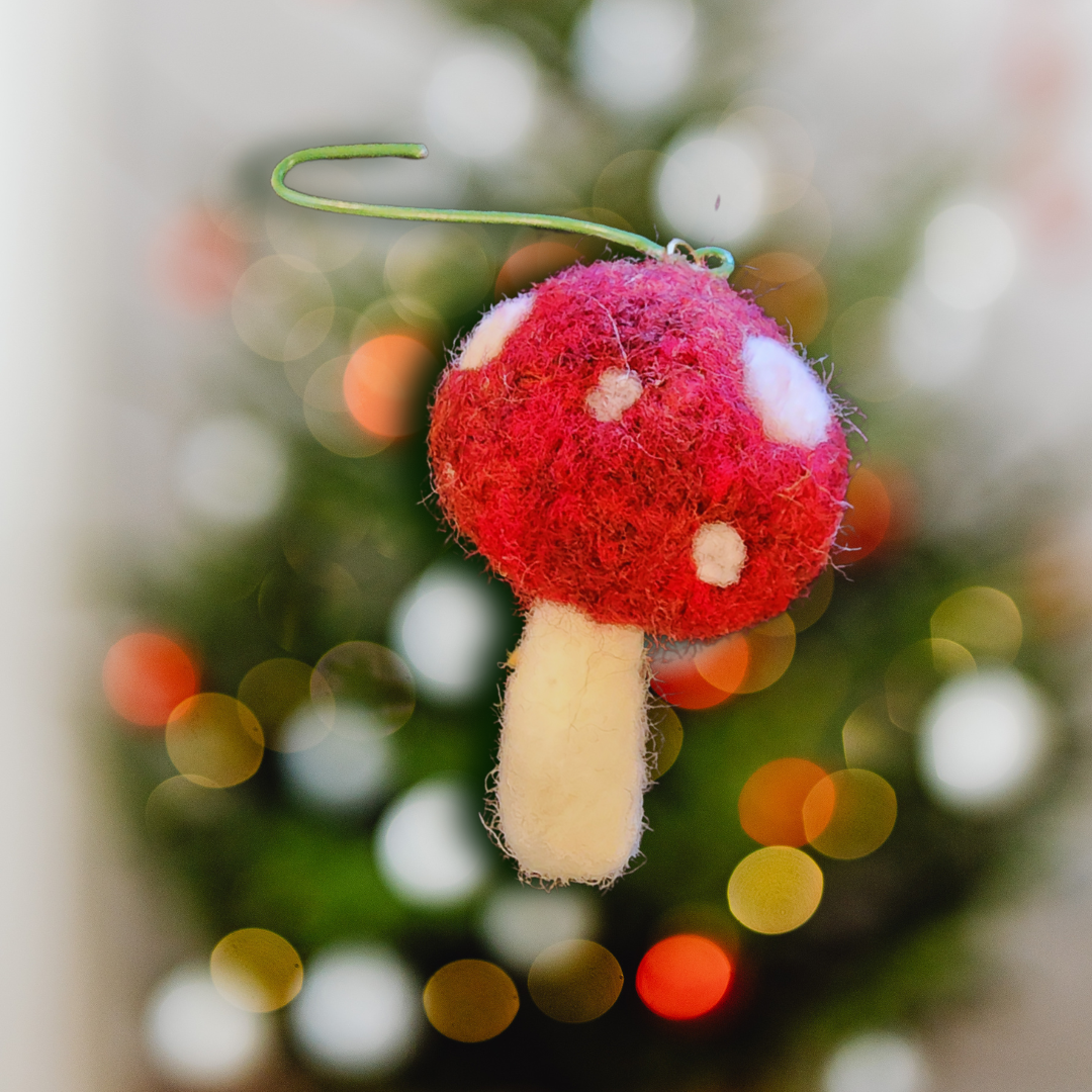 A needle-felted red and white toadstool ornament hangs from a green hook against a blurry background of a bright Christmas tree with holiday lights (bokeh effect). This image highlights the mushroom's classic shape.