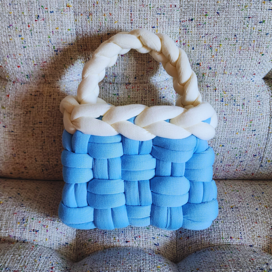 A small handmade basket weave handbag, featuring a light blue woven body and a thick white braided handle, sitting upright on a multicolored speckled fabric surface.