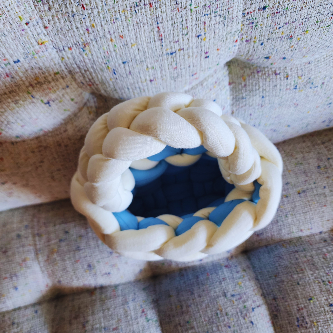 An overhead view looking down into a small handmade basket weave handbag, showing its light blue interior and the white braided top edge/handle, resting on a textured fabric surface.