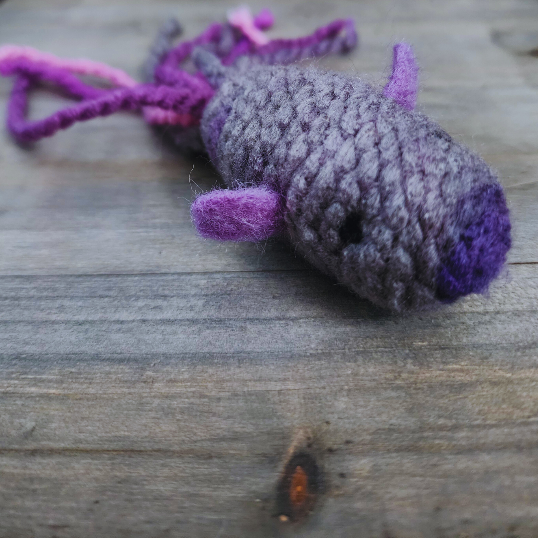 A close-up, slightly angled view of a handmade catnip fish cat toy, with a textured gray and purple body, a small black eye, and purple fins, resting on a wooden background.