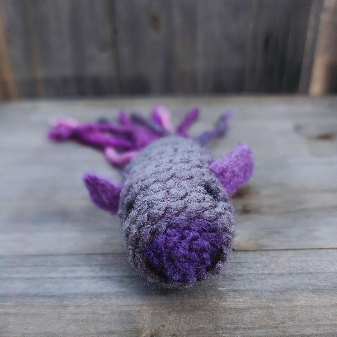 A close-up front view of a handmade catnip fish cat toy, showing its gray body fading to purple at the nose, with purple fins and a blurred multi-colored purple tail in the background, on wood.