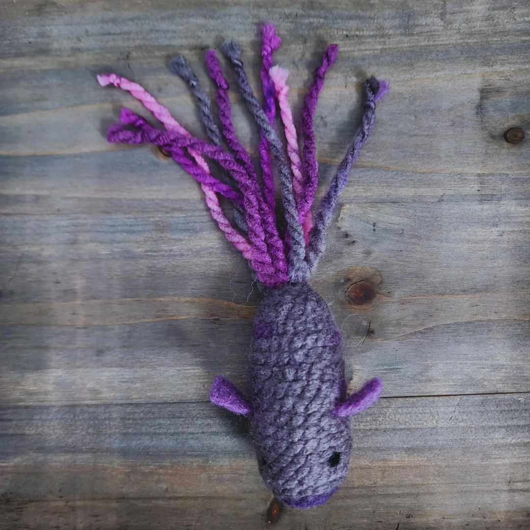 A handmade catnip fish cat toy in shades of gray and purple, with a long, tentacled tail of various purple and pink yarn strands, lying flat on a wooden surface.
