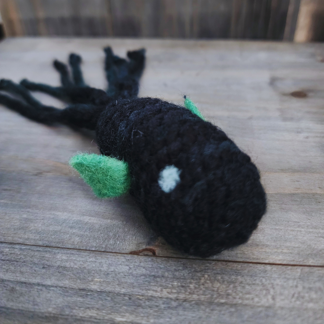 A handmade catnip fish cat toy in dark multi-colored yarn with green fins and a light blue spot, on a wooden surface.