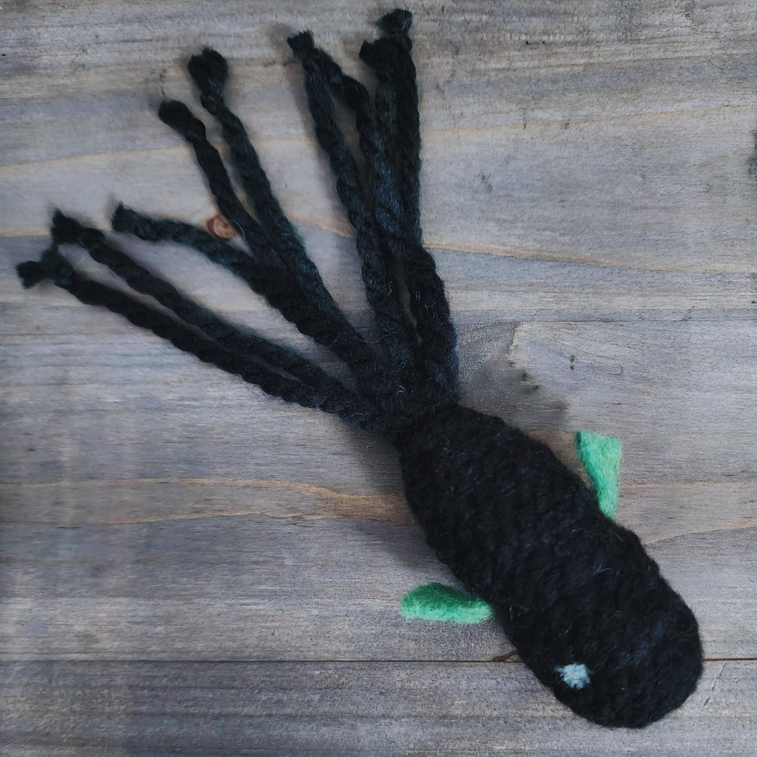 A handmade catnip fish cat toy in dark multi-colored yarn with green fins, a light blue spot, and a long, dark tentacled tail, lying on a wooden surface.