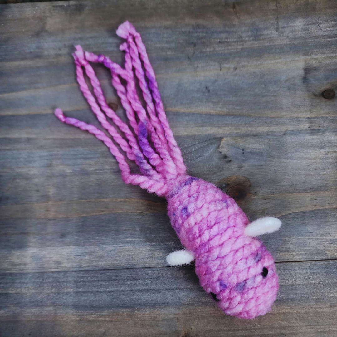 A handmade catnip fish cat toy in mottled pink and purple yarn with a long, tentacled tail and small white fins, lying flat on a wooden surface.
