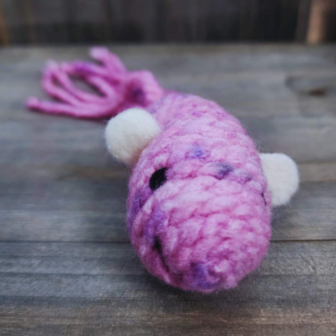 A close-up view of the head of a handmade catnip fish cat toy, showing its mottled pink and purple yarn body, black eye, and small white fin, against a wooden background.