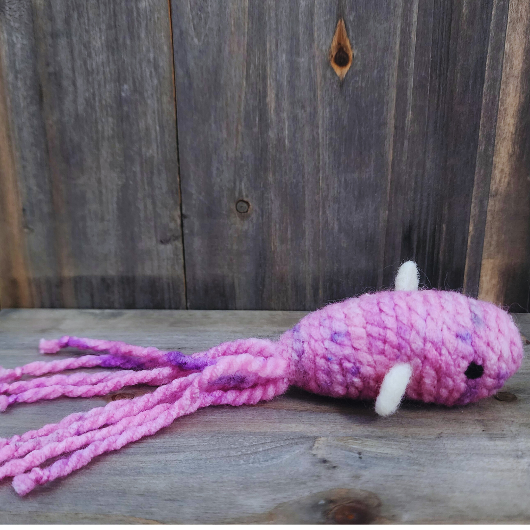 A handmade catnip fish cat toy in mottled pink and purple yarn with a long, tentacled tail and small white fins, lying stretched out on a wooden surface.