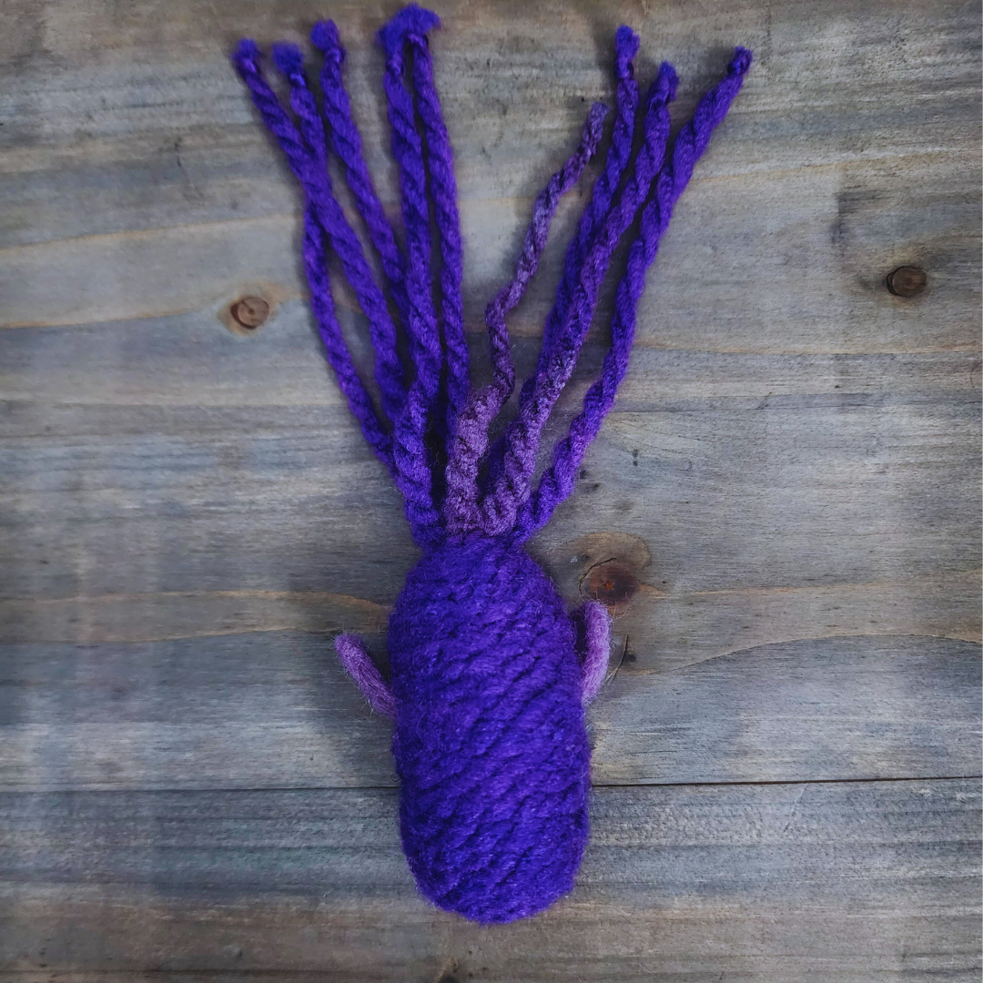 A handmade catnip fish cat toy in vibrant purple yarn, with a long, tentacled tail of various purple shades, lying flat on a wooden surface.