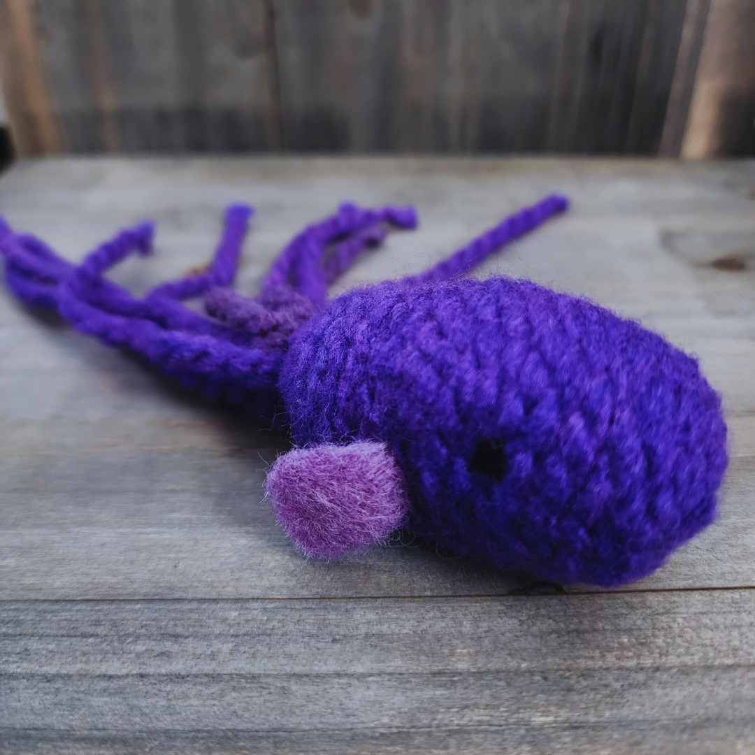 A close-up view of a handmade catnip fish cat toy in vibrant purple, showing its textured body, small black eye, and lighter purple fin, resting on a wooden background.