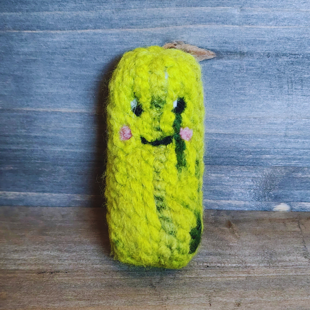 A handmade, bright green pickle cat toy standing upright against a rustic blue wood background. The toy features a needle-felted kawaii face with small pink cheeks and a smiling expression.