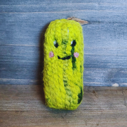 A handmade, bright green pickle cat toy standing upright against a rustic blue wood background. The toy features a needle-felted kawaii face with small pink cheeks and a smiling expression.