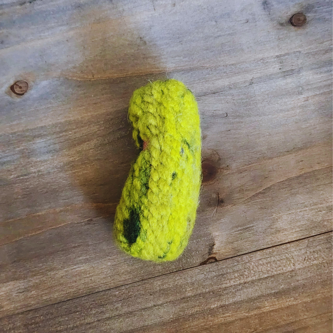 A side view of a handmade green felted pickle cat toy resting on a natural wood surface, showing its thick, durable texture and slightly curved shape.