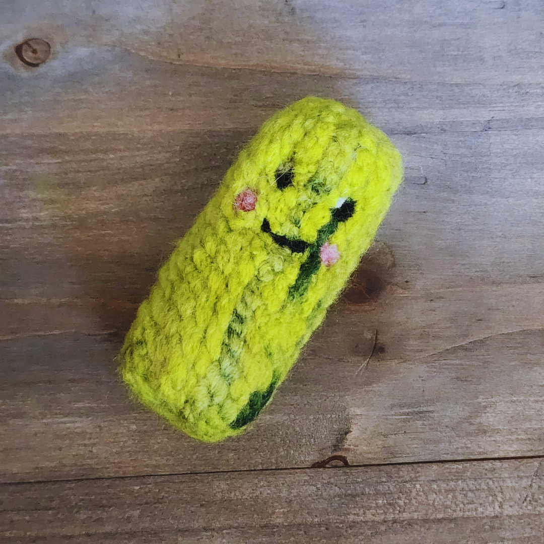 A handmade catnip pickle toy lying diagonally on a wooden surface. The green textured body is accented with dark green "pickle" spots and a hand-felted smiling face.
