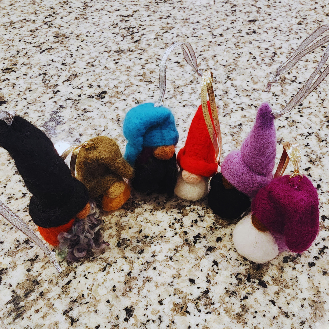 A row of six whimsical needle-felted gnome ornaments with various colored hats (red, blue, purple, brown, black), beards, and silver or gold ribbon hangers, resting on a speckled granite countertop.