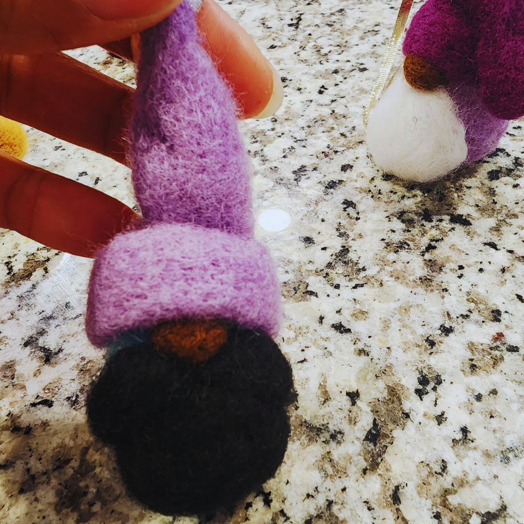 Close-up of a hand holding a gnome ornament with a lavender-purple conical hat and a black beard.