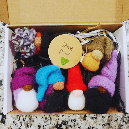 Overhead view of a set of five brightly colored, needle-felted gnome ornaments packaged in an open cardboard box with black shredded paper filling. A circular "Thank You" tag is visible in the center.