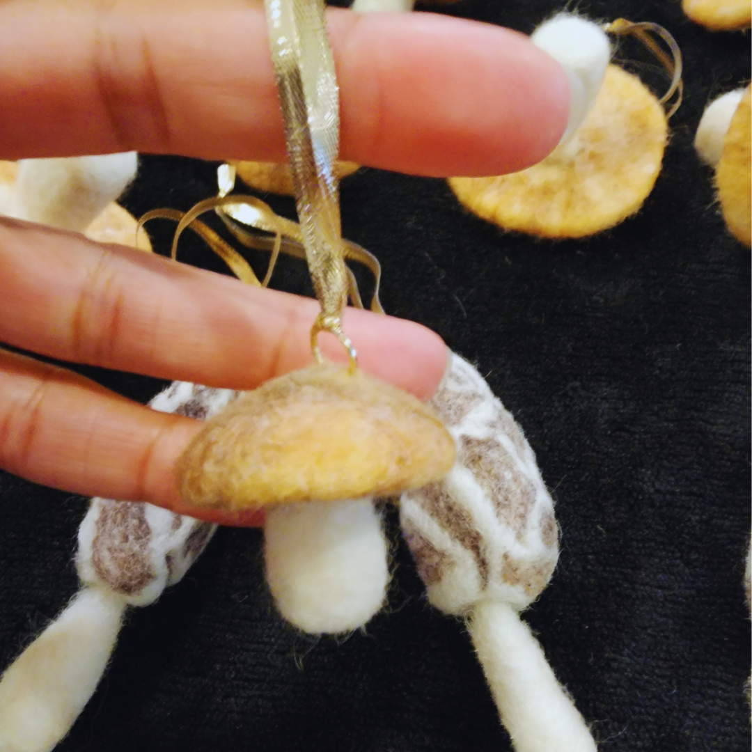 Close-up of a hand holding one round, classic-shaped needle-felted mushroom ornament with a soft orange cap, white stem, and gold ribbon hanger.